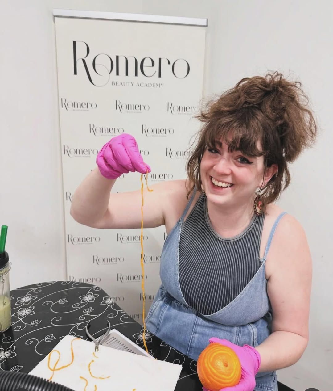 Hands-on demo in pink gloves in front of the Romero Beauty Academy backdrop